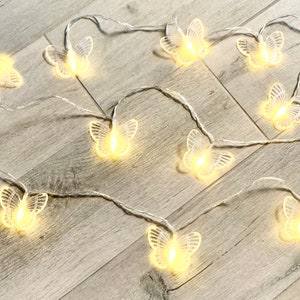 Può includere: Una ghirlanda di luci con lampadine trasparenti a forma di farfalla. Le luci sono infilate su un filo bianco e sono accese, proiettando un bagliore caldo.