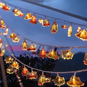 May include: String of fairy lights with various camping-themed designs, including tents, campfires, and lanterns. The lights are strung on a white wire and are illuminated with warm white bulbs.