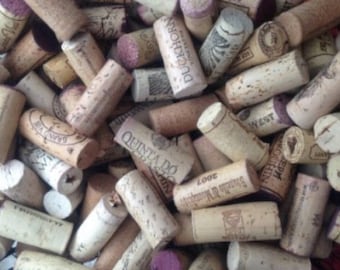 May include: A large collection of cylindrical natural cork wine stoppers in various shades of tan and brown. Many of the corks feature printed text and logos, including brands like Duckhorn, Quinta do Noval, and Brunello di Montalcino.