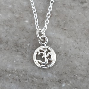 May include: A silver-toned necklace featuring a small, circular pendant with an openwork Om symbol design. The pendant is suspended from a delicate, thin silver-toned chain with small, uniform oval links that connect to a jump ring at the top.