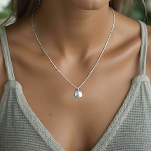 Peut inclure: Un collier à chaîne en argent délicat avec un petit pendentif rond en argent poli. Le collier repose sur un haut sans manches gris clair texturé avec une encolure en V. Le métal présente une finition brillante et réfléchissante.