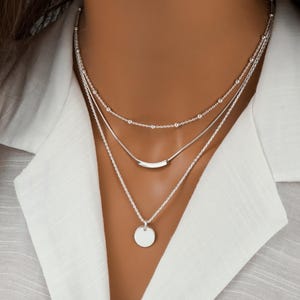 May include: A three-layered silver necklace featuring a delicate beaded chain, a smooth curved bar pendant on a medium chain, and a small circular disc pendant on the longest chain. The necklace is worn against a white collared shirt.