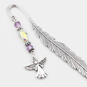 May include: A silver-toned metal bookmark shaped like a feather. Attached to the top loop is a dangling charm featuring two small purple faceted beads, one iridescent clear oval bead, and a small silver-toned metal angel figure at the end.