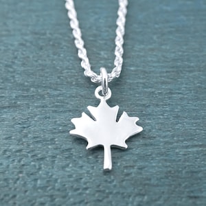 May include: A silver-colored necklace featuring a small, flat maple leaf pendant suspended from a twisted rope chain. The pendant has a smooth, polished finish and is attached to the chain by a simple circular jump ring bail.