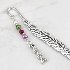 May include: A silver-toned metal bookmark shaped like a feather with a dangling charm attachment. The charm features a vertical row of four small round beads in white, green, red, and purple, followed by a silver-toned DNA double helix charm.