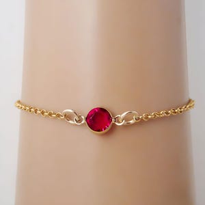 May include: A delicate gold chain bracelet featuring a single, round, faceted ruby-red gemstone set in a polished gold bezel. The bracelet is constructed with small, uniform circular links and secures with a simple, integrated metal clasp mechanism.