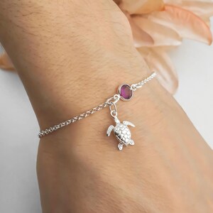 May include: A silver chain bracelet featuring a small, detailed sea turtle charm and a single round, deep pink gemstone set in a silver bezel. The bracelet is delicate, with a polished finish that reflects light, creating a subtle and elegant look.