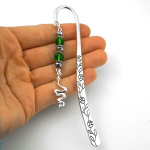 May include: A silver-toned metal hook bookmark featuring a floral vine engraving along the stem. A dangling charm attachment consists of two faceted emerald green beads, two smaller dark grey beads, and a silver-toned snake-shaped metal charm.
