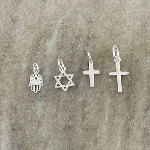 May include: A set of four silver-toned metal charms featuring a Hamsa hand, a Star of David, and two different cross designs. Each charm includes a small circular jump ring at the top for attachment, displaying a polished, reflective metallic finish.