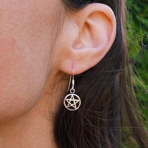 May include: A silver earring featuring a pentagram charm. The drop earring has a small hook for the ear. The charm is a five-pointed star enclosed within a circular frame, a symbol often associated with Wicca.