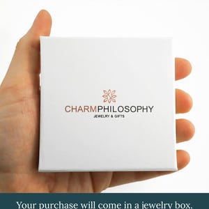 A hand holding a white jewelry box with the text "CHARMPhilosophy JEWELRY & GIFTS" printed on it.