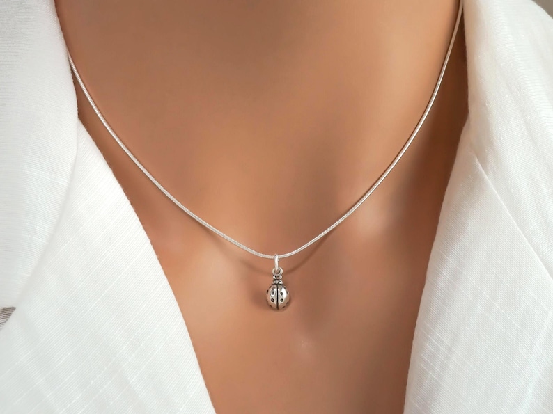 Puede incluir: Un collar de plata con un peque&ntilde;o colgante de mariquita detallado suspendido de una cadena de plata fina y lisa. El dije de mariquita tiene puntos negros visibles y un acabado met&aacute;lico pulido sobre una prenda blanca.