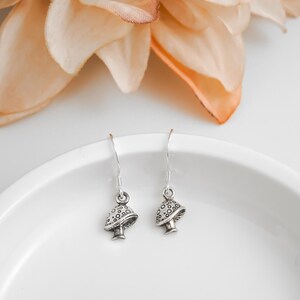 May include: A pair of silver mushroom earrings with detailed caps and stems. The earrings are displayed on a white plate, with a peach-colored flower in the background. The earrings are a whimsical accessory.