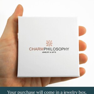 A hand holding a white jewelry box with the text "CHARMPhilosophy JEWELRY & GIFTS" printed on it.