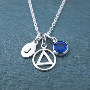 May include: A silver-toned necklace featuring three charms suspended from a delicate chain. The charms include a small oval disc engraved with the letter J, a circular pendant with an open triangle cutout, and a round, faceted deep blue crystal.