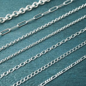 May include: A selection of silver chain necklaces in different styles, including round, rectangular, and oval links. The chains are displayed against a teal background, highlighting their varied patterns and textures.
