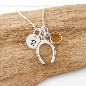 Peut inclure: Un collier argenté composé d'une chaîne fine avec trois breloques : un petit disque circulaire gravé de la lettre R, une breloque fer à cheval à la texture martelée et une pierre précieuse ronde à facettes de couleur ambre sertie d'argent.