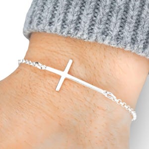 May include: A silver bracelet featuring a cross design. The bracelet is made of a delicate chain with a cross pendant. The cross is centered on the wrist, and the bracelet is secured with a clasp.