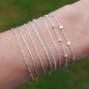 May include: An assortment of silver bracelets in various chain styles, including figaro, snake, and beaded designs. The bracelets are displayed on a person's arm against a blurred green background, showcasing the different textures and patterns.