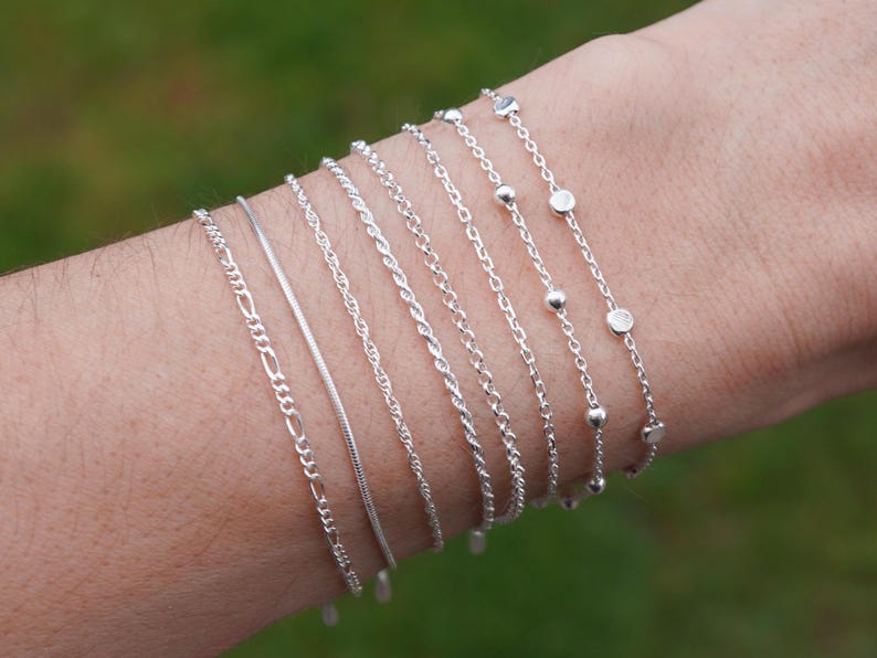 May include: An assortment of silver bracelets in various chain styles, including figaro, snake, and beaded designs. The bracelets are displayed on a person's arm against a blurred green background, showcasing the different textures and patterns.