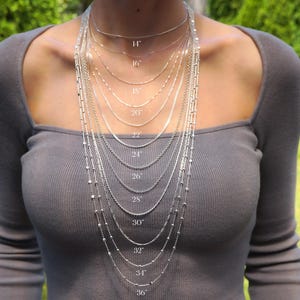 a woman wearing a necklace with multiple chains of varying lengths, each labeled with a specific measurement in inches. The necklace appears to be a chain length chart, which helps determine how the chain fits based on the wearer's height and neck circumference.