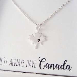 May include: A silver necklace featuring a maple leaf pendant with a tiny heart. The necklace is presented on a white card, with the words "WE'LL ALWAYS HAVE Canada" in a handwritten font.