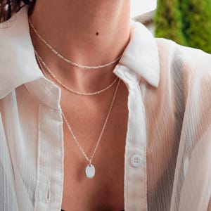 May include: A layered silver necklace set consisting of two delicate chains and a third longer chain featuring a small, smooth, oval-shaped pendant. The jewelry is worn over a white button-down shirt with a visible collar and white buttons.