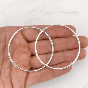 May include: A pair of large, thin, circular hoop earrings crafted from polished silver-toned metal. The earrings feature a simple, continuous wire design with a smooth, reflective surface and a seamless closure mechanism at the top of each hoop.