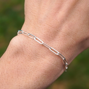 May include: A silver link bracelet is shown on a wrist. The bracelet is composed of small, rectangular links. The background is a blurred green, suggesting an outdoor setting. The bracelet is a simple, elegant piece of jewellery.