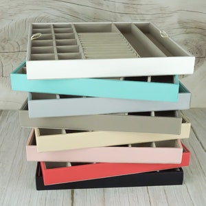 Peut inclure: Une pile de plateaux à bijoux colorés dans diverses nuances, dont blanc, turquoise, gris, beige, rose, corail et noir. Chaque plateau a un intérieur gris clair avec des compartiments pour organiser les bijoux. Les plateaux sont empilés les uns sur les autres.