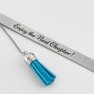 May include: A silver metal bookmark with the text "Enjoy the Next Chapter!" and a turquoise tassel. The bookmark has a chain connecting the tassel to the main body. The tassel is made of a soft, fringed material.
