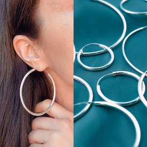 May include: A collection of polished silver hoop earrings in various sizes. The hoops are circular, thin, and metallic with a reflective finish. One hoop is shown worn in an earlobe, while several other hoops of differing diameters are arranged together.