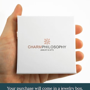 A hand holding a white jewelry box with the text "CHARMPhilosophy JEWELRY & GIFTS" printed on it.