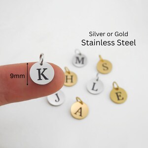 May include: Round silver and gold stainless steel charms, each featuring a single engraved letter. The charms measure 9mm in diameter. The text Silver or Gold Stainless Steel is visible. The letters K, M, S, H, L, J, A, and E are displayed.