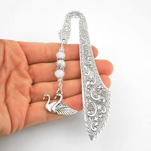 May include: Silver-toned bookmark featuring a swan charm and decorative floral etching. The bookmark has a curved top and a dangling strand of white beads. The swan charm is detailed with feathers. A unique accessory for book lovers.