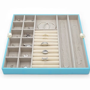 May include: A square jewellery organiser tray with a bright turquoise exterior and a beige velvet interior. The tray features multiple compartments, including small square sections, a ring holder section with horizontal slots, and two long rectangular channels for necklaces and hoop earrings.