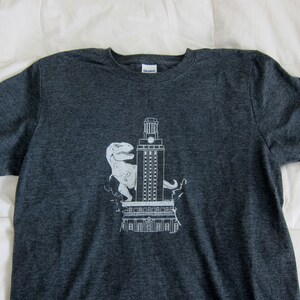 May include: A dark gray t-shirt with a white graphic of a dinosaur standing on top of the University of Texas Tower.