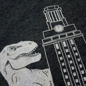 May include: A black t-shirt with a white graphic print of a dinosaur and a building with a clock tower.