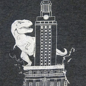 May include: A white graphic print on a dark grey t-shirt shows a Tyrannosaurus Rex dinosaur standing on top of the University of Texas at Austin's iconic tower.