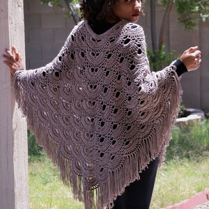 Puede incluir: Un poncho de ganchillo color topo con un dobladillo con flecos. El poncho presenta un diseño calado detallado con un patrón de vieira. La prenda se usa sobre una camiseta y pantalones negros, creando un look en capas. La imagen está tomada al aire libre.