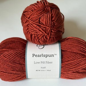 Op de afbeelding: Drie bollen roestkleurig garen zijn op een wit oppervlak gestapeld. De onderste twee bollen liggen naast elkaar, met een label met de tekst "Pearlspun Low Pill Fiber rust". De bovenste bol rust op de andere twee. Elke bol weegt ongeveer 100 gram.
