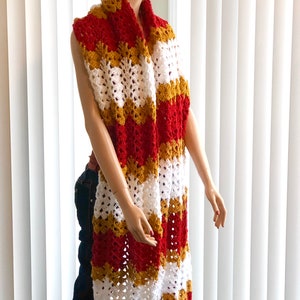 May include: A long crocheted scarf in red, white, and gold stripes. The scarf is draped around a mannequin's neck.