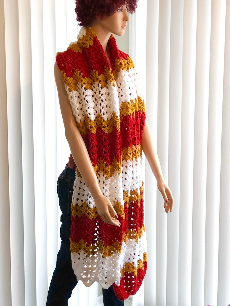 May include: A long crocheted scarf in red, white, and gold stripes. The scarf is draped around a mannequin's neck.