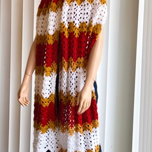 May include: A long, crocheted scarf in red, white, and gold stripes. The scarf is made with a simple stitch pattern and has a fringed edge.