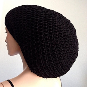 May include: A black, oversized crochet beanie hat. The hat is made of a textured, woven material and has a slouchy fit. The hat is shown on a mannequin head, with the back of the head and neck visible.