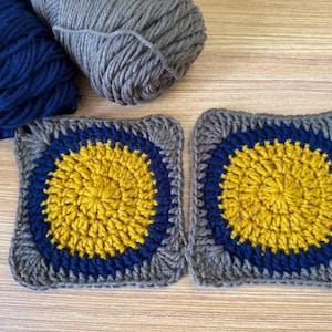 May include: Two crocheted squares with a yellow centre, surrounded by a blue ring and a grey border. Two balls of yarn, one navy blue and one grey, are in the background. The squares are handmade and likely for a craft project.