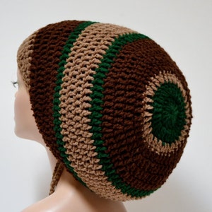 May include: A handmade crochet hat in brown, beige, and green. The hat features a circular design with alternating bands of colour. The hat is shown on a mannequin head, showcasing its shape and texture. The hat has a drawstring.