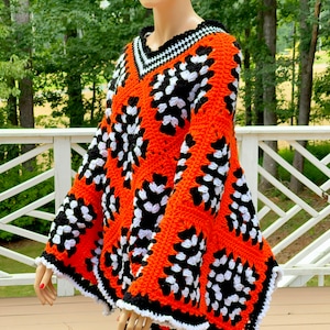 Puede incluir: Un poncho de ganchillo con un patrón geométrico en naranja, negro y blanco. El poncho tiene cuello en V y mangas acampanadas.