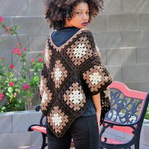 Puede incluir: Un poncho de ganchillo marrón, beige y blanco con un patrón de cuadrados de la abuela. El poncho lo lleva una persona con el pelo rizado y se combina con pantalones negros.