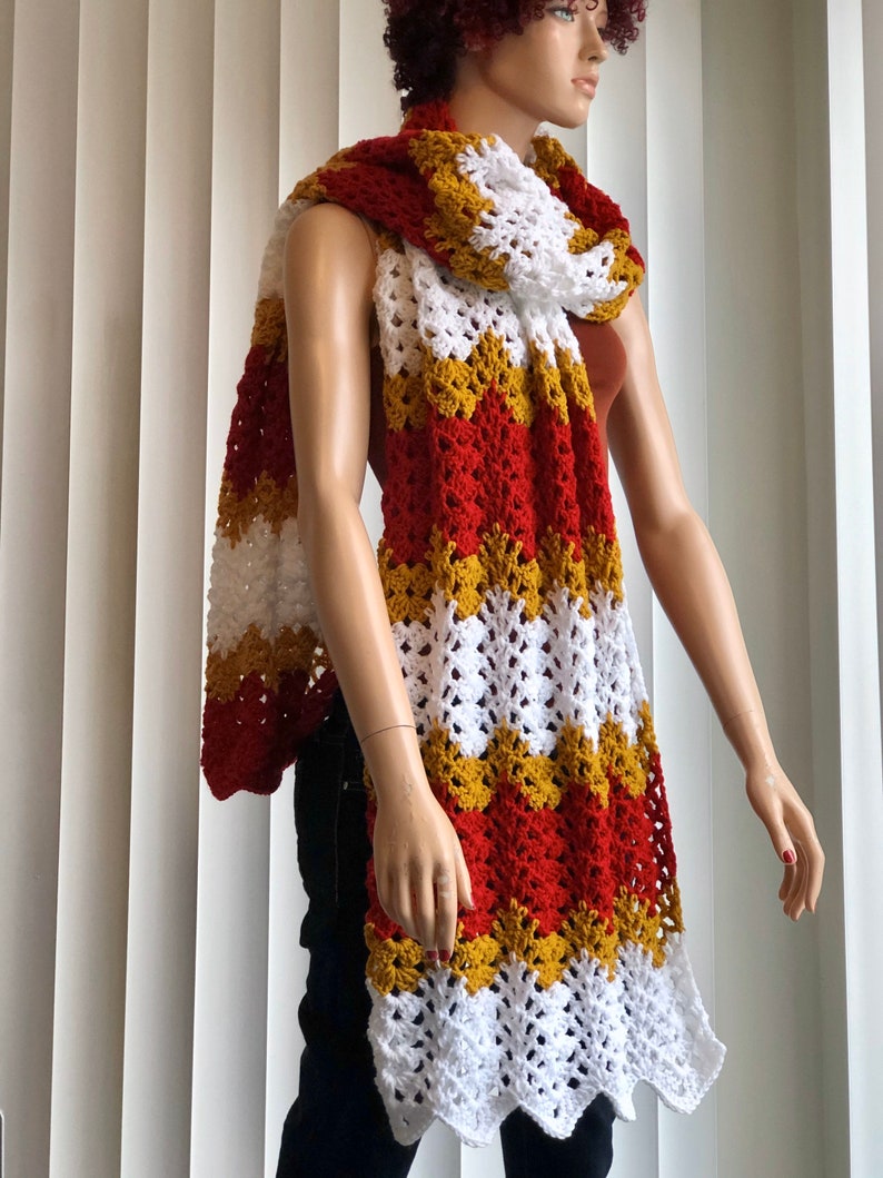 May include: A long, crocheted scarf with red, white, and gold stripes. The scarf is made with a lacy stitch and has a scalloped edge.